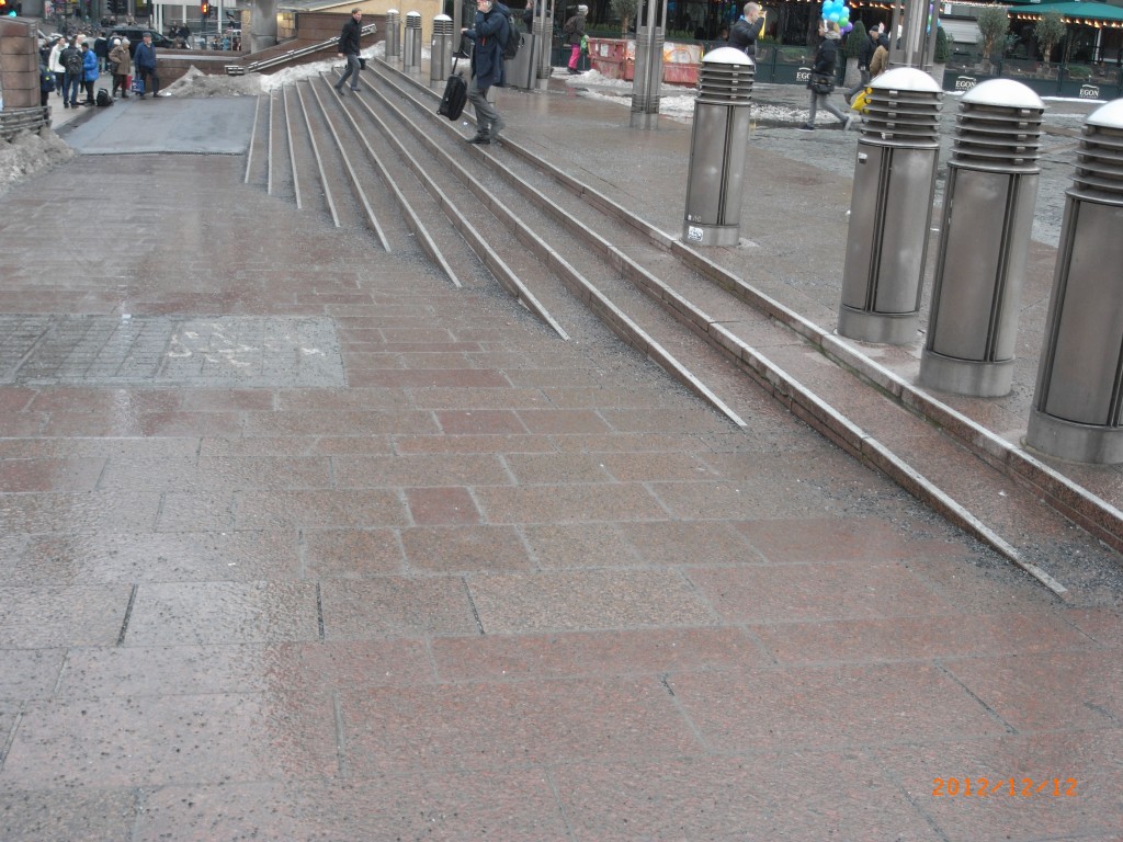 Image shows the approach to an Oslo train station. In the background are shallow stairs, marked with reflective borders and textured grip. In the foreground is a shallow ramp to the same level